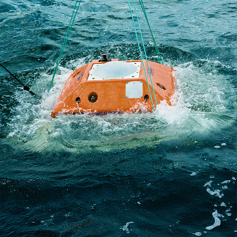 Orange oceanographic instrument suspended by ropes being lowered into choppy water.
