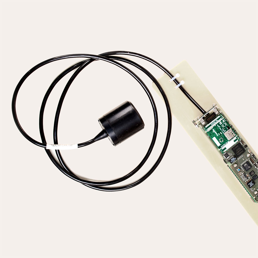 Electronic circuit board connected to a black cylindrical sensor with coiled black cable against a light background.