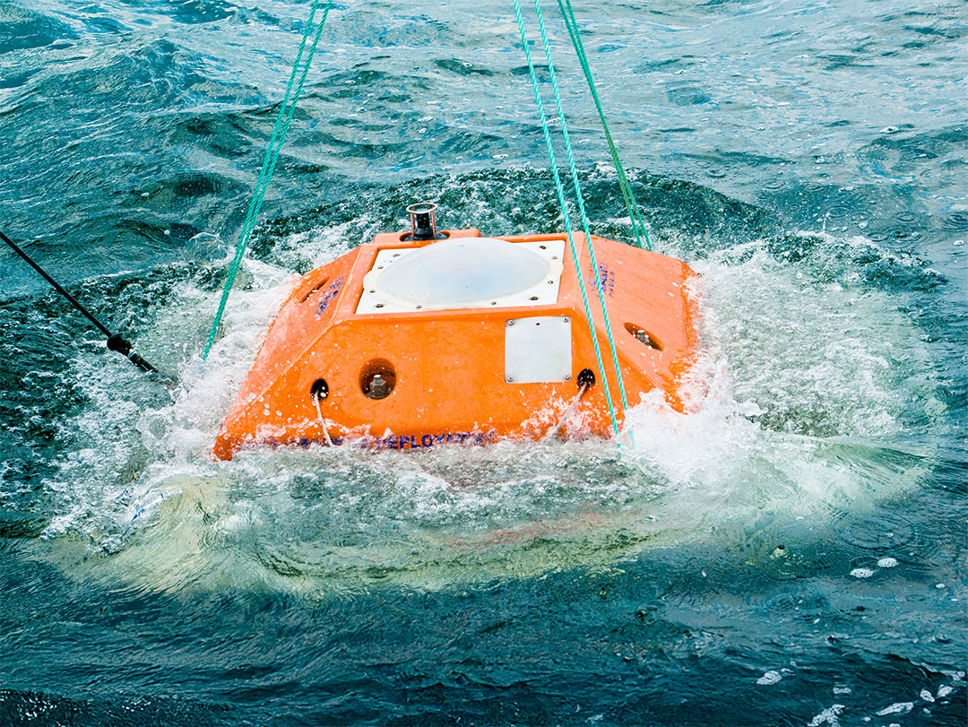 Orange marine instrument or equipment being lowered into or lifted from the water with green ropes attached.