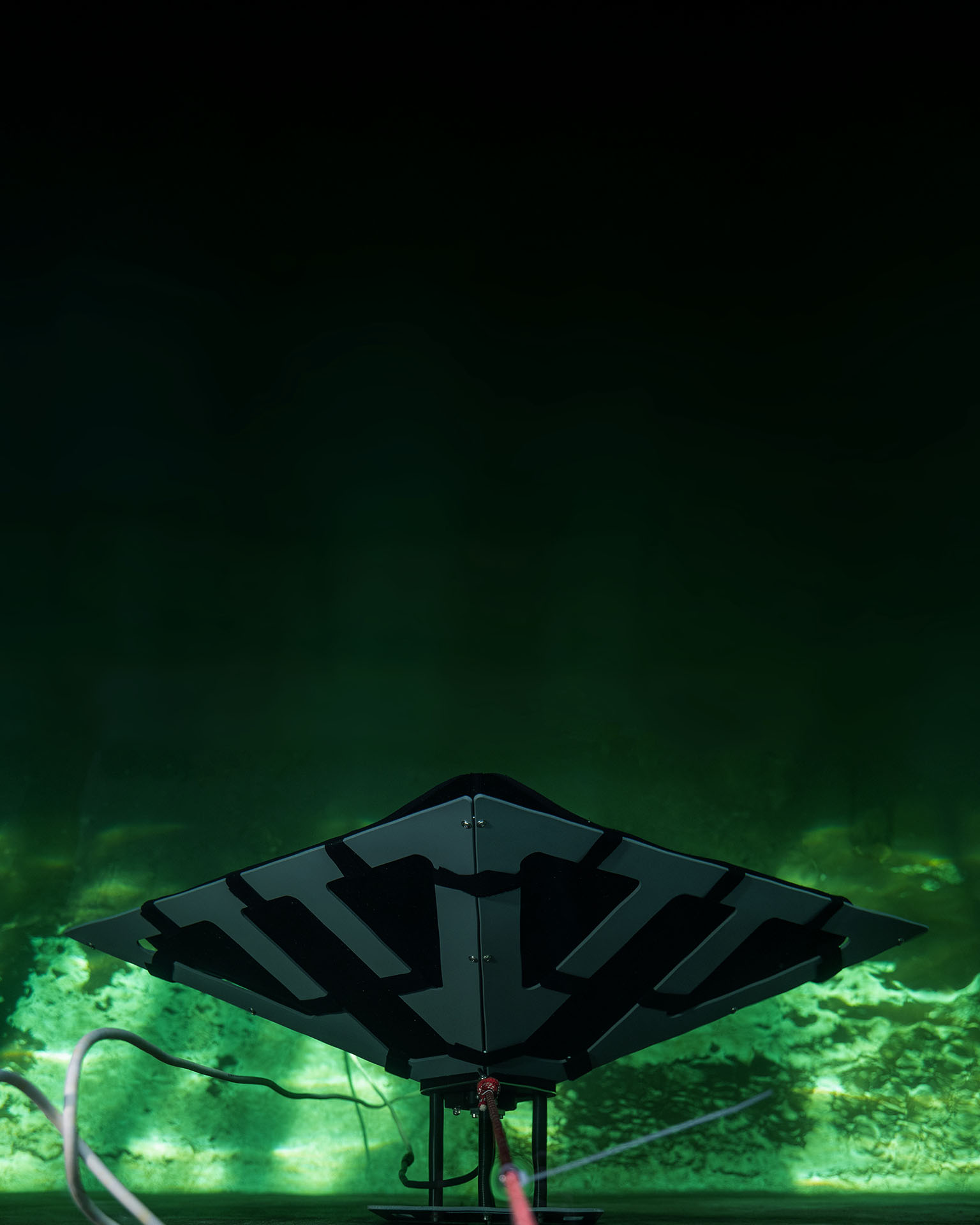 Underwater view of a black and gray flat rectangular device tethered with cables against a green aquatic background.