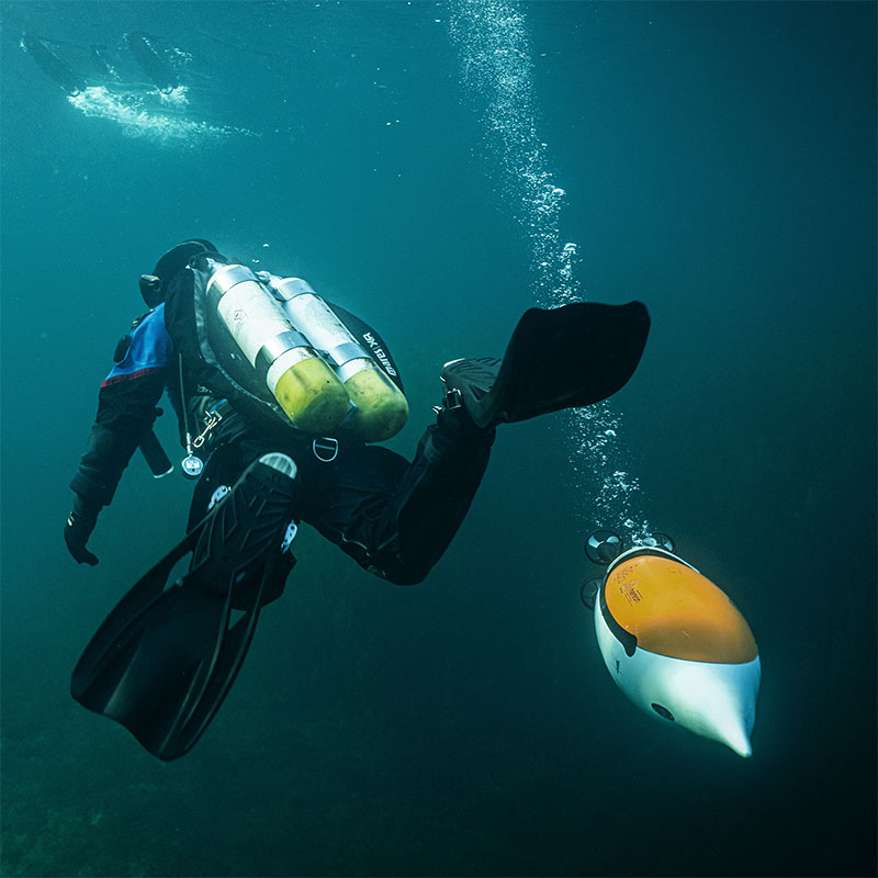 Scuba diver underwater swimming alongside a small yellow and white underwater drone releasing bubbles.