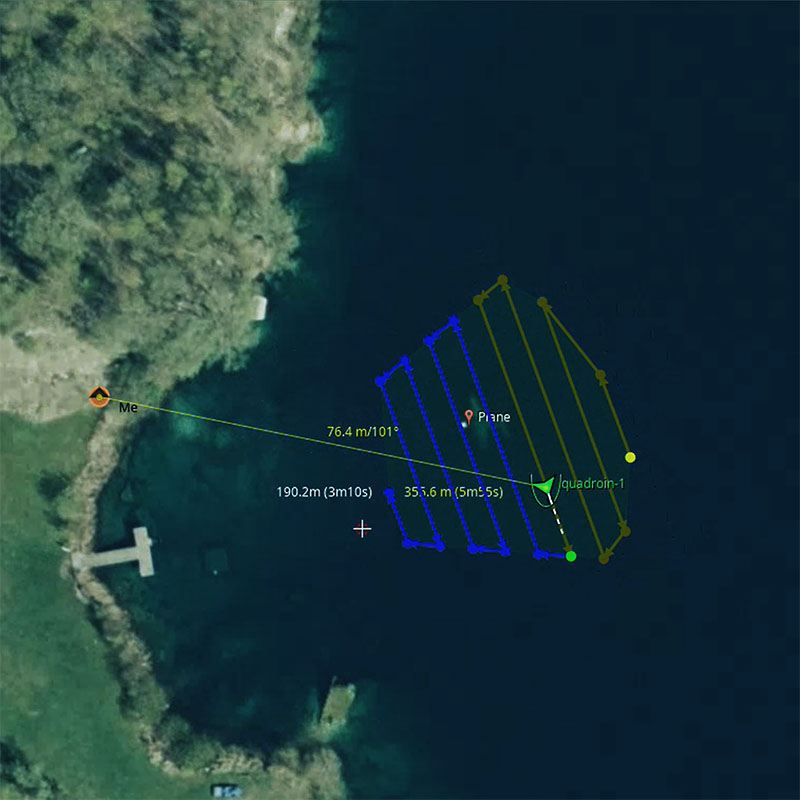 Aerial map view showing drone flight paths over a body of water near a wooded shoreline, with distance and time metrics, and labeled points for 'Me,' 'Plane,' and 'quadroin-1'.
