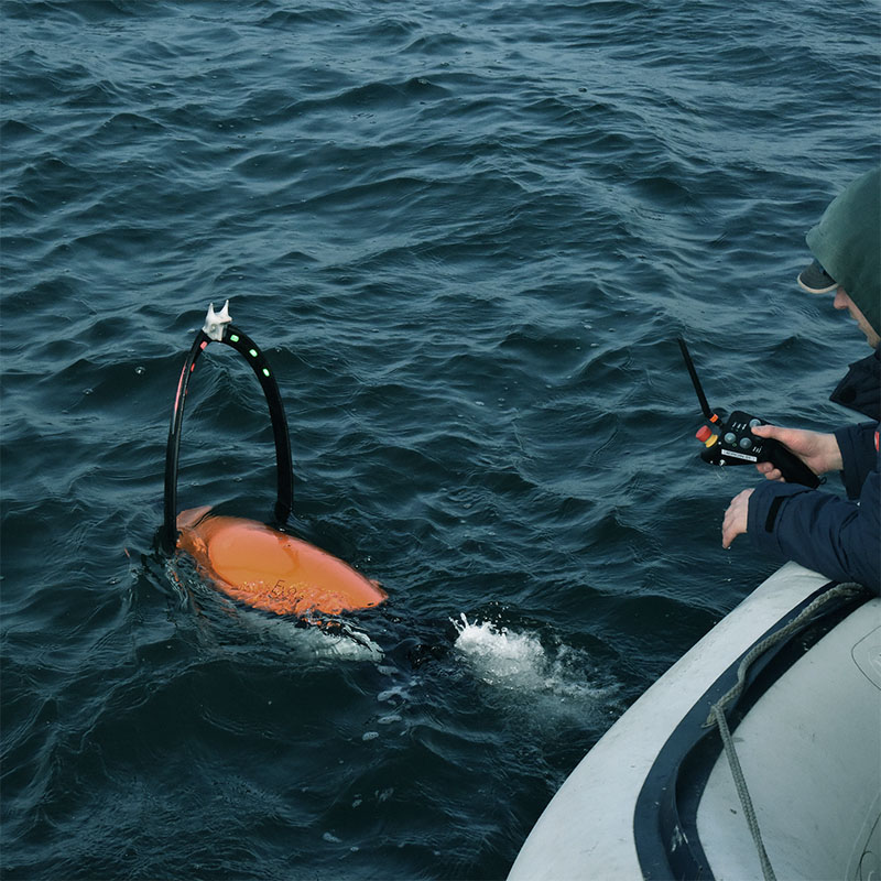 Person on a boat operating a remote-controlled orange underwater drone in dark, choppy water.