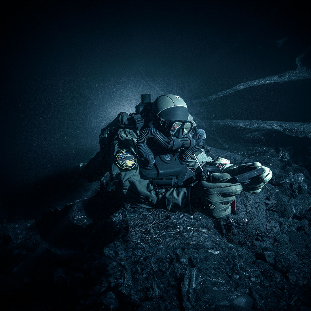 Scuba diver in full gear exploring a dark underwater environment, illuminated from behind.