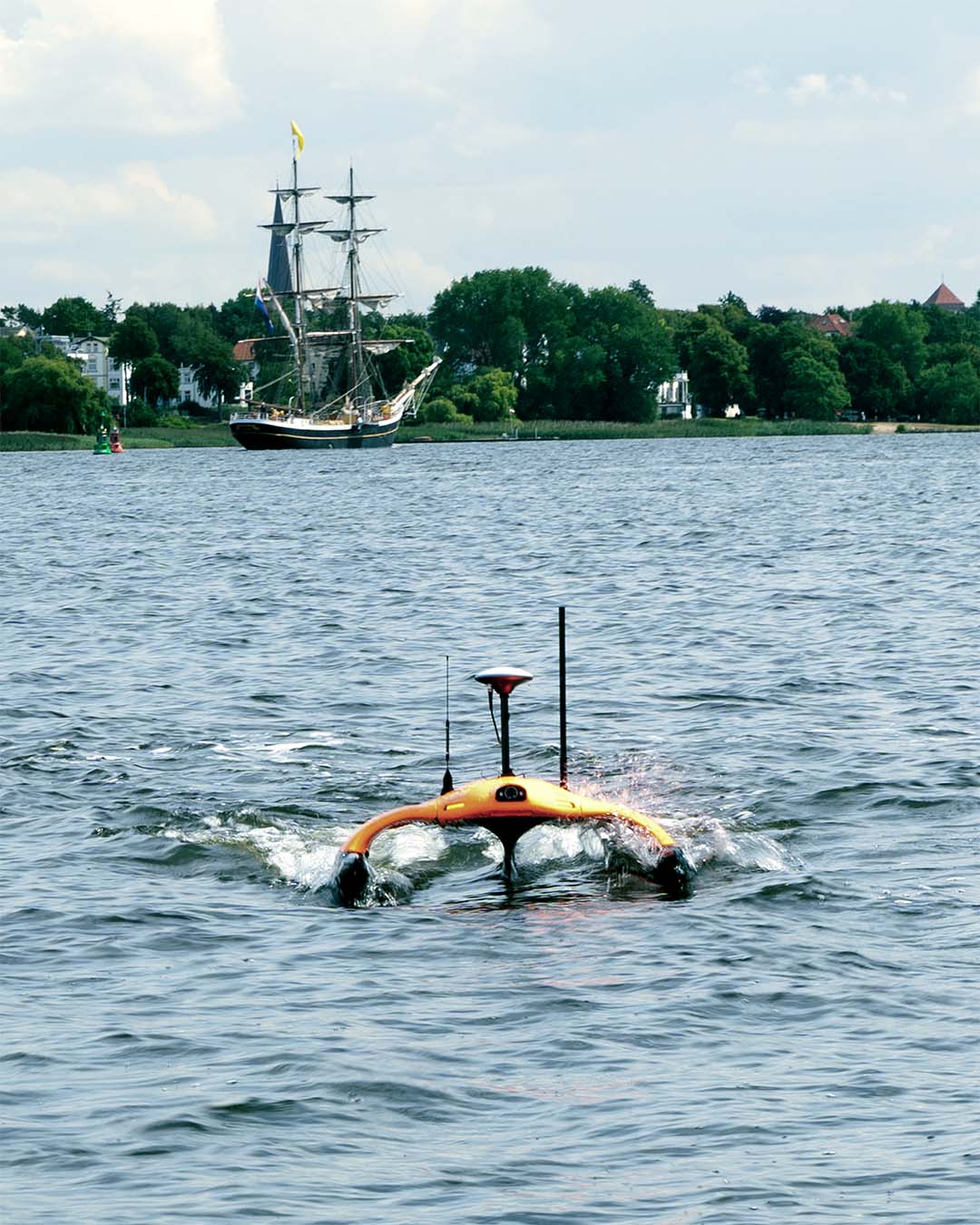 Orange autonomous water drone moving on a lake with a sailing ship and tree-lined shore in the background.