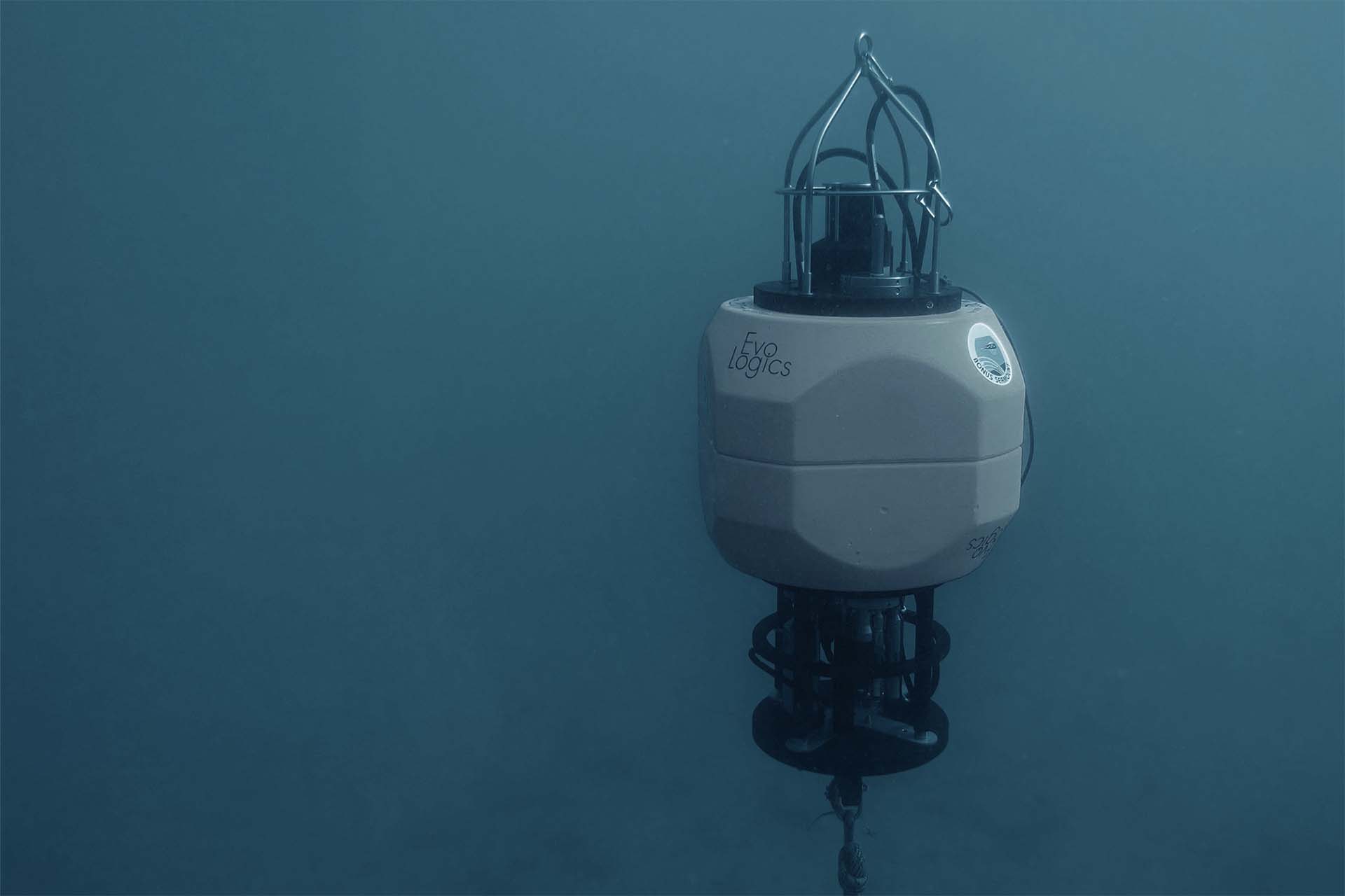 Underwater scientific instrument labeled EvoLogics suspended in water with attached cables and metal frame.