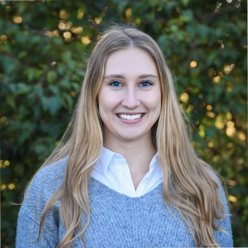 Smiling woman with long blonde hair wearing a gray sweater and white collared shirt in front of green foliage.
