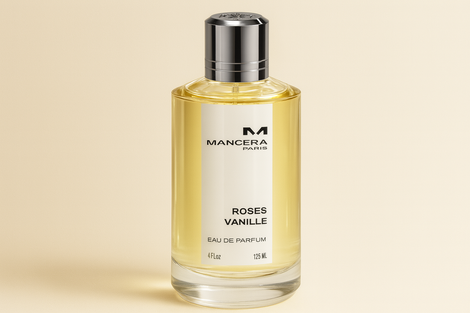 A realistic widescreen image of Mancera Roses Vanille Eau de Parfum bottle with light golden liquid, silver cap, and black text label, set against a soft cream gradient background.