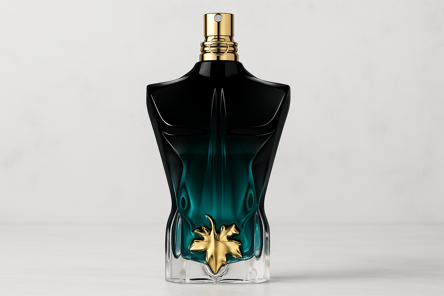  A realistic widescreen image of Jean Paul Gaultier Le Beau Le Parfum bottle with a black-to-teal gradient torso design, gold leaf emblem, and gold atomiser, set against a light marble-textured background.