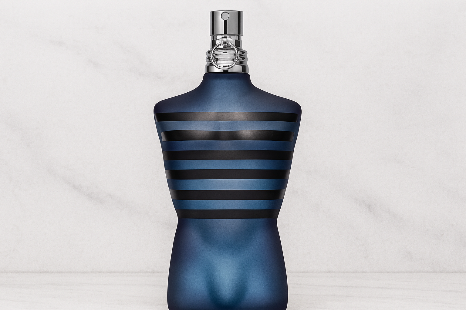 A realistic widescreen image of Jean Paul Gaultier Ultra Male perfume bottle shaped like a deep blue torso with black stripes and a silver atomiser, set against a light marble-textured background.
