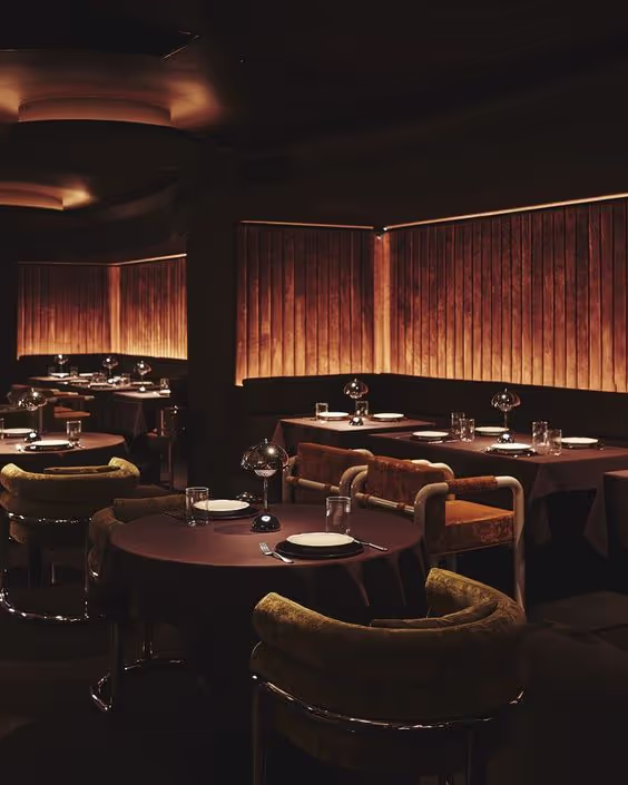 Dimly lit restaurant interior with round tables, cushioned chairs, and wooden paneling with warm ambient lighting.