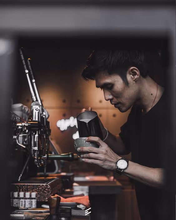Barista with a watch pouring steamed milk into a cup at a coffee machine in a dimly lit café.