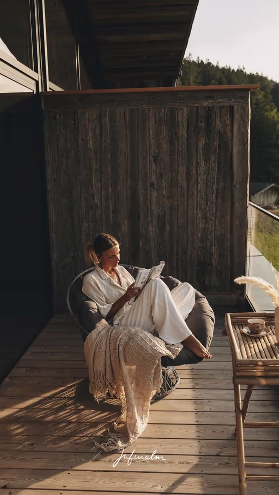 Woman in white loungewear reading a newspaper while sitting on a cushioned chair on a wooden balcony with a blanket and small table holding a cup.