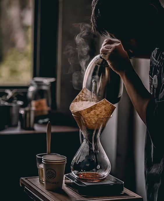 Person pouring hot water from a kettle into a coffee filter on a glass coffee maker with steam rising.