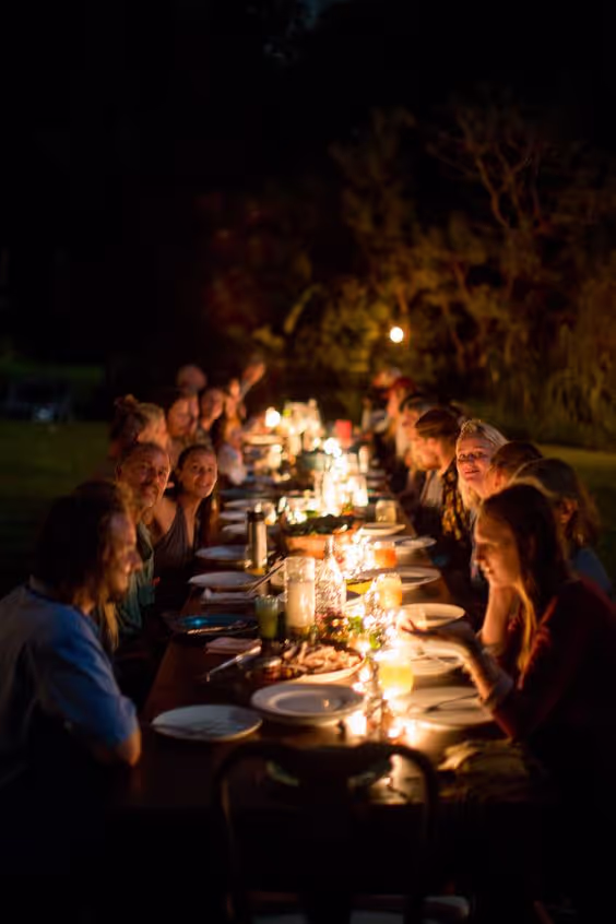 Group of people gathered around a long outdoor table lit by candles at night, sharing a meal.