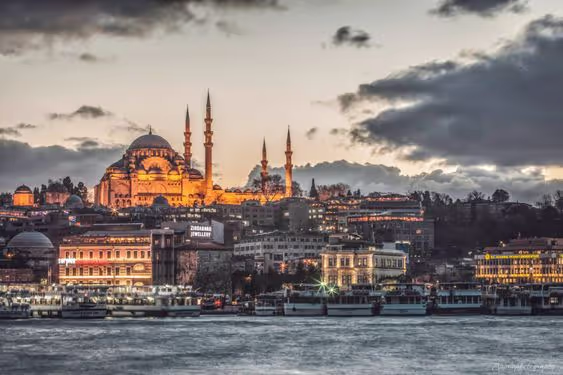 Evening view of illuminated Suleymaniye Mosque with multiple minarets overlooking buildings and boats on the water in Istanbul.