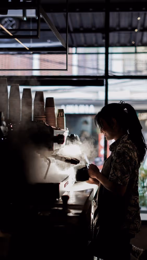 Barista steaming milk with espresso machine in a coffee shop with light streaming through large windows.