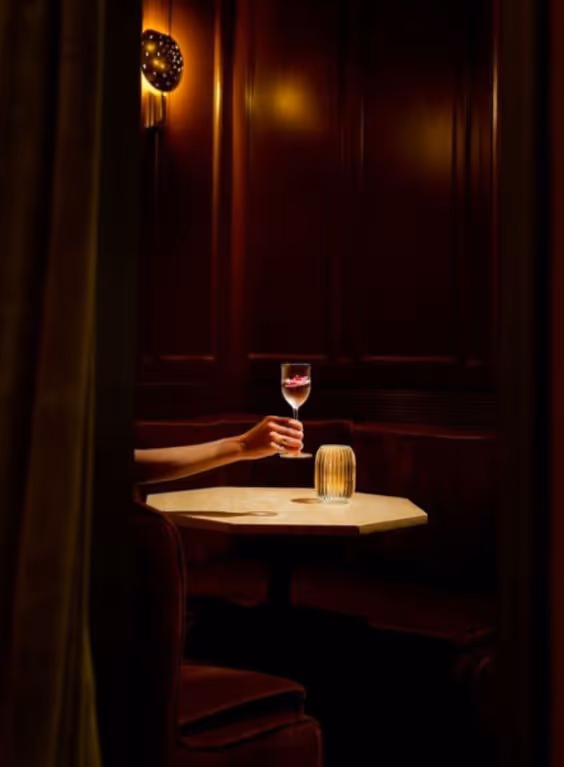A hand holding a wine glass with red liquid over a round table in a dimly lit, cozy seating area with dark wood paneling and warm lighting.