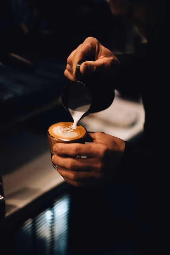 Barista pouring steamed milk to create latte art in a glass cup with a dark background.