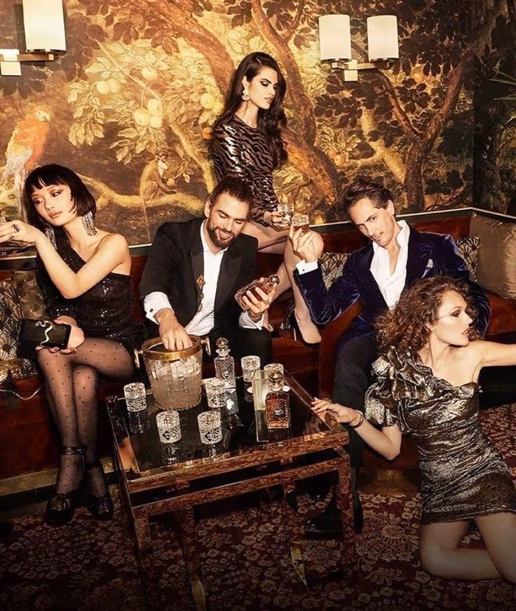 Five stylishly dressed people socializing in a vintage-style lounge with a decorative wall mural, glassware, and bottles on a table.