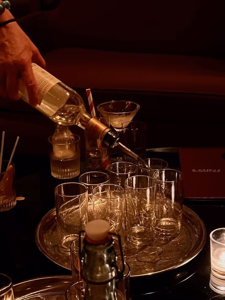 A hand pours clear liquid from a bottle into one of several empty glasses on a round tray in dim lighting.
