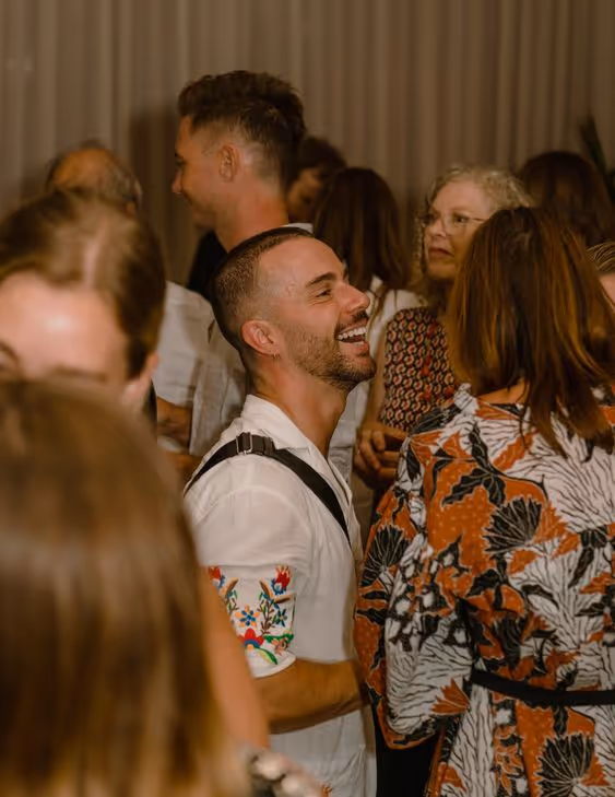 A man smiling and laughing in a crowded room with people conversing around him.