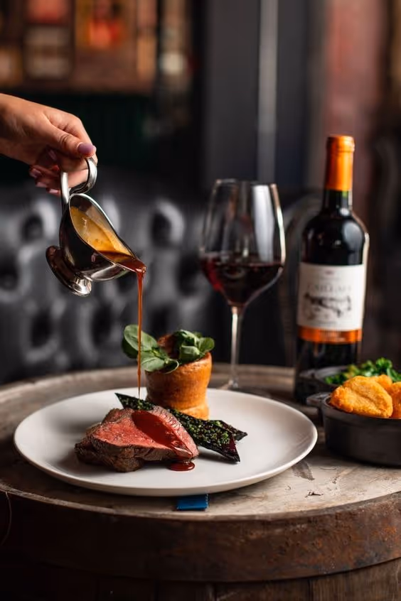 Hand pouring sauce over sliced medium-rare steak on a plate with greens and a baked side, accompanied by a glass and bottle of red wine.