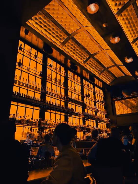 Silhouettes of people sitting and standing at a bar with backlit shelves filled with bottles under a patterned, illuminated ceiling.