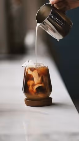 Cream being poured from a metal pitcher into a glass of iced coffee on a coaster.