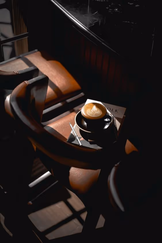 Black cup of latte with leaf latte art on a saucer, placed on a chair with leather seating in dim light.