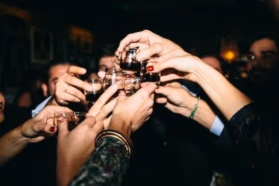 Group of people clinking shot glasses filled with dark liquid in a celebratory toast.