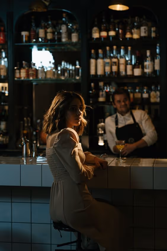 Woman in a beige dress sitting at a tiled bar counter with a bartender behind the bar filled with bottles.