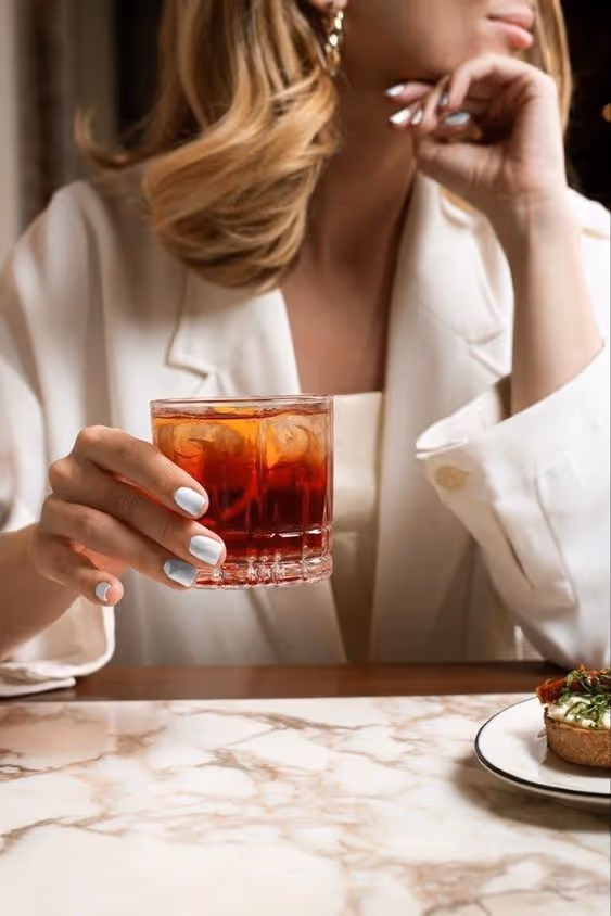 Woman in cream blazer holding a glass with an iced red drink at a marble table next to a plate with a small appetizer.