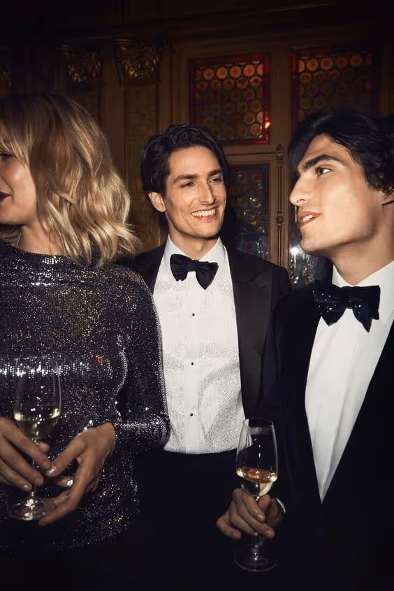 Two men in tuxedos and bow ties and a woman in a sparkly dress holding wine glasses at a formal event.