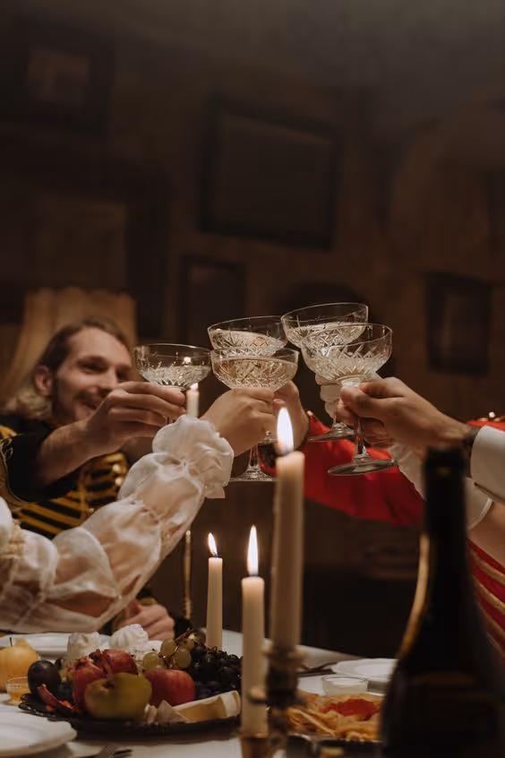 Four people clinking crystal champagne glasses in a warm, candlelit setting with fruit and food on the table.