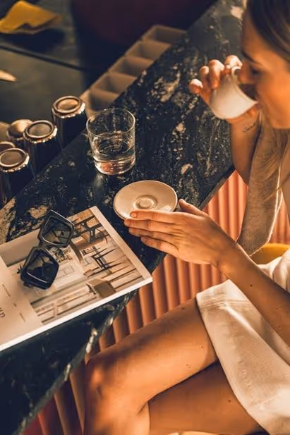 Woman sitting at a marble countertop drinking from a small cup with sunglasses and a magazine placed on the counter nearby.