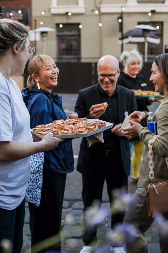 Group of people smiling and enjoying small pastries outdoors, with one person holding a tray full of pastries.