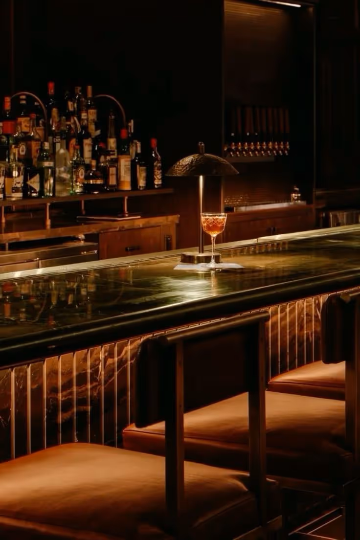 Dimly lit bar countertop with cushioned stools, a cocktail glass under a small lamp, and various liquor bottles in the background.