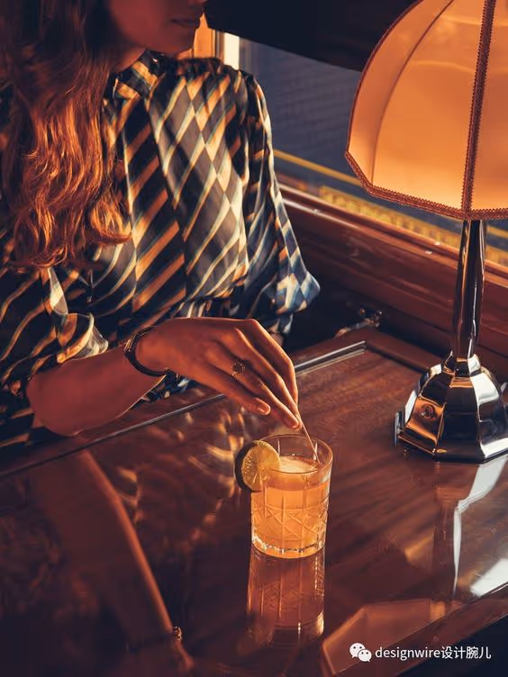 Woman in a striped blouse stirring a drink with a lime garnish at a wooden table beside a lamp.