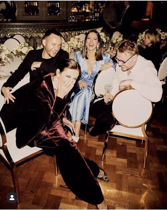 Four people sitting and laughing around a table at an indoor event with wooden flooring and floral decorations.
