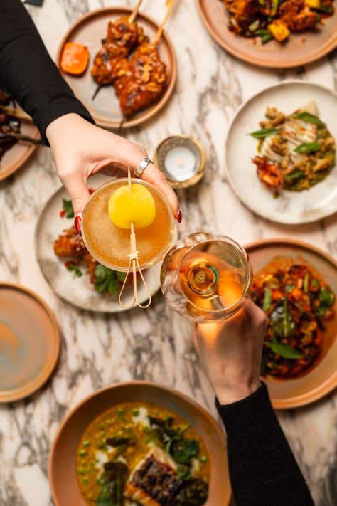 Two hands holding cocktails above a marble table set with various dishes of food.