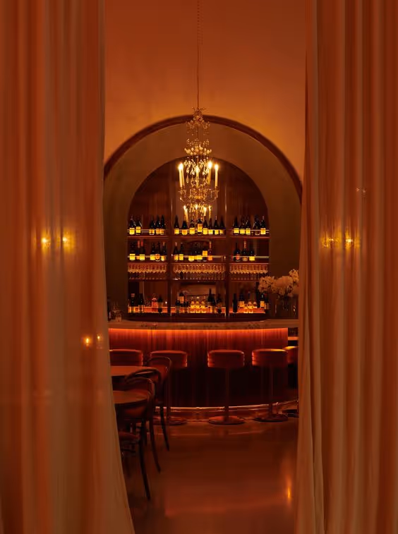 Warmly lit bar with a chandelier, stocked shelves of bottles, and bar stools visible through open curtains.