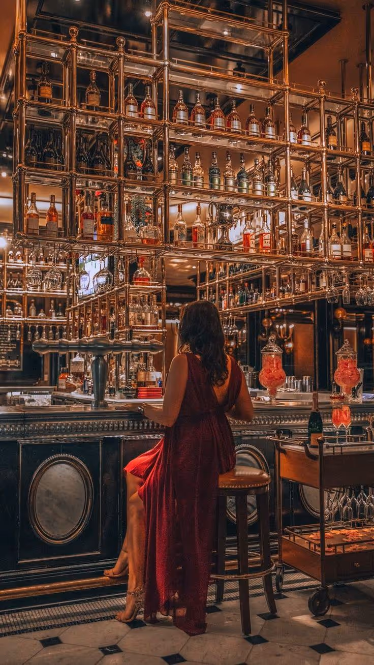 Woman in a red dress sitting on a bar stool facing a luxurious bar stocked with various bottles of alcohol and glassware.