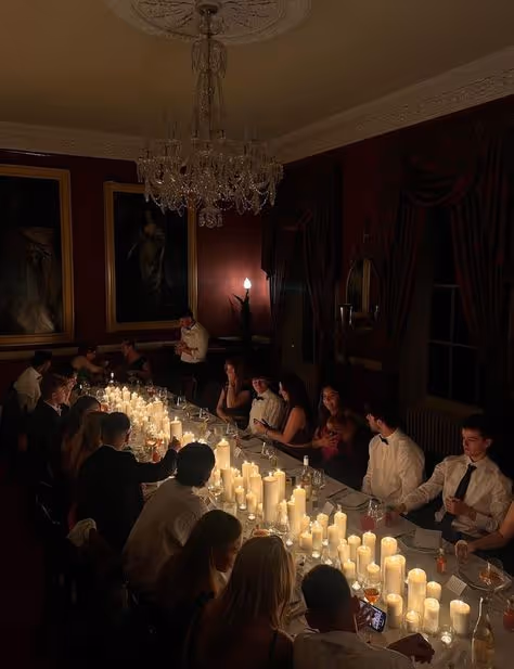 A dimly lit dinner party with people seated around a long table illuminated by numerous candles.