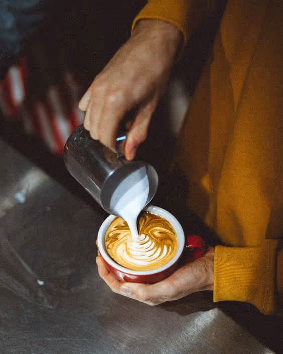 Person pouring steamed milk into a red cup of coffee to create latte art.