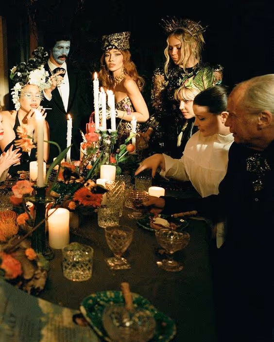 A group of elegantly dressed people gathered around a candlelit dinner table adorned with flowers and crystal glassware in a dimly lit setting.