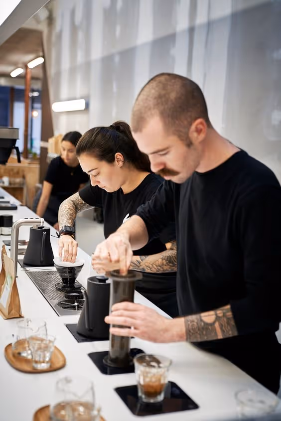 Two baristas concentrating as they prepare coffee using pour-over and AeroPress methods on a white counter in a modern cafe.