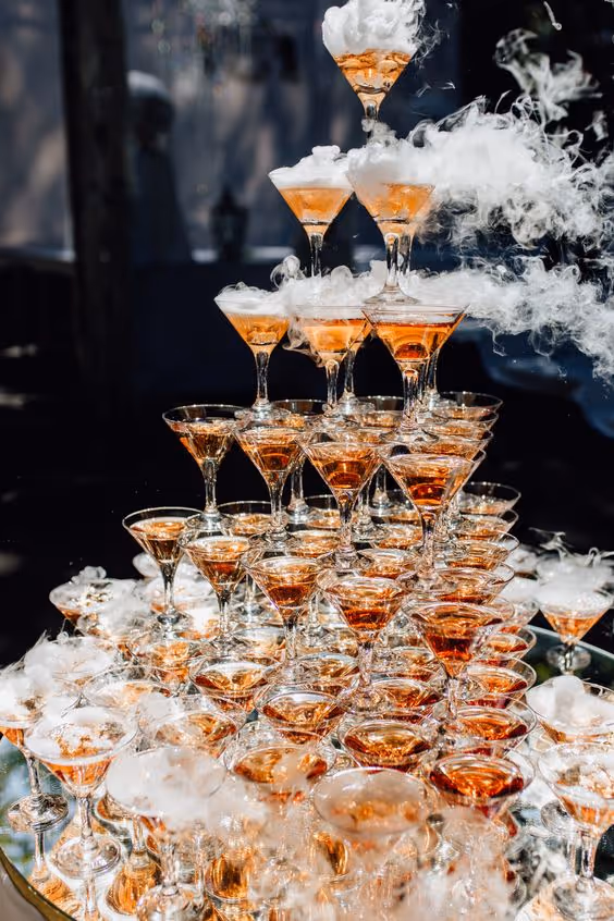 Pyramid stack of champagne glasses filled with amber liquid, surrounded by white smoke or fog effects.