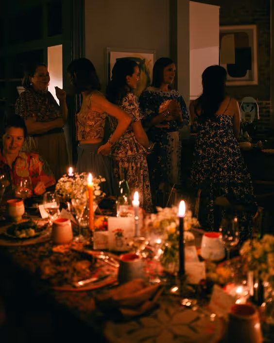 Dimly lit dinner party with women standing and chatting near a table set with candles, flowers, and food.
