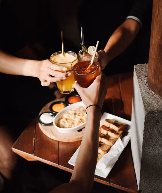 Three people clinking cocktails over a wooden table with a bowl of dip and sliced toasted bread.